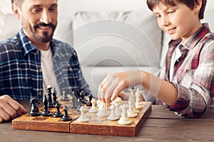 Father and little son at home sitting at table playing chess moving pieces happy close-up blurred