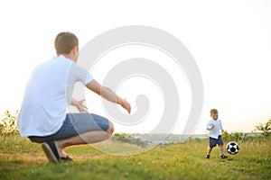 father with a little son plays football on the green grass in the park. Happy family having fun and playing football