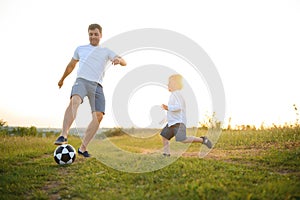 father with a little son plays football on the green grass in the park. Happy family having fun and playing football