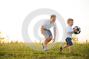 father with a little son plays football on the green grass in the park. Happy family having fun and playing football