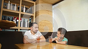 Father and little son sitting in cafe with juice and drinking