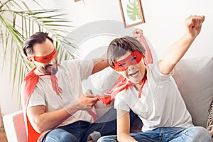 Father and son in superheroe costumes at home sitting on sofa boy close-up hand up shouting motivated while dad holding photo