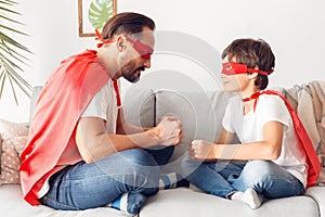 Father and son in superheroe costumes at home sitting on sofa clencing fists playing looking at each other joyful side photo