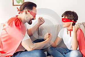 Father and son in superheroe costumes at home sitting on sofa holding hands looking at each other excited team spirit photo
