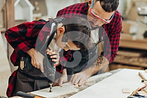 Father and little son working with drill perforating wood plank at workshop