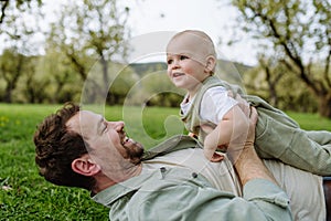 Father lying and playing with little baby, having fun in grass during warm spring day. Father& x27;s day concept.
