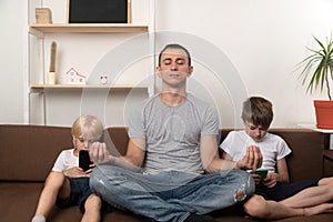 Father meditates while children sit and look at phone. Dad has calmed his sons and is doing yoga