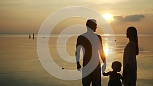 Father, mother and daughter stand on tropical beach look at sunset. Dad and mum keep daughter`s hand. slow motion