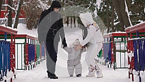 Father and mother with her son walk down an alley in a winter park holding baby hands. Mom corrects a pacifier to his