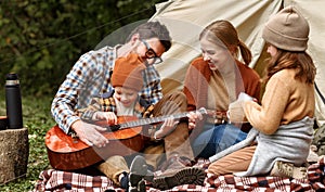 Father, mother and little son sitting near tourist tent and playing guitar during camping