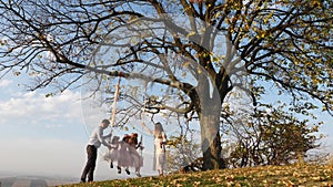 Father and mother shakes her daughters on a swing under a tree.