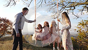 Father and mother shakes her daughters on a swing under a tree.