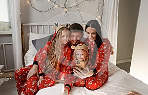 Father, mother and two little daughters in bed are photographed looking at the phone. Happy family in red Christmas