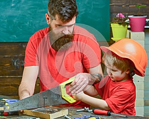 Father, parent with beard teaching little son to sawing with sharp handsaw, carpenter crafts. Boy, child busy in