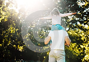 Father and playful daughter from behind having fun in sun at park with copyspace on green trees. Loving parent carrying
