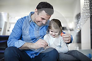 Father playing with little daughter sitting on couch and helping to paint her nails with bright polish play together
