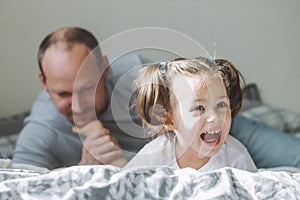 Father plays with little daughter 2-4 on bed. Dad tickles kids feet with his beard. Family, having fun