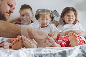 Father plays with three children on bed. Dad tickles kids feet. Family of daddy, two girls, and boy