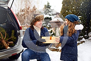 Father presents daughter a gift box on snowy winter day outdoors. Christmas tree in large trunk of family car. Girl