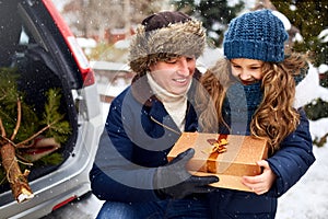 Father presents daughter a gift box on snowy winter day outdoors. Christmas tree in large trunk of family car. Girl