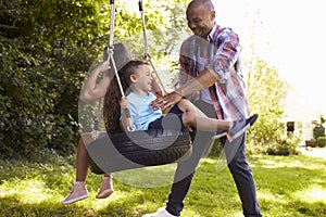 Father Pushing Children On Tire Swing In Garden