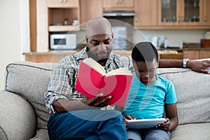 Father reading book while son sitting next to him using digital tablet