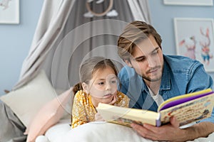 Father reading a book to his daughter while lying on the floor in bedroom.