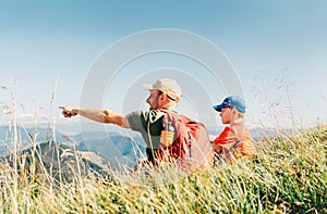 Father showing something interesting to his teenager son sitting on the grass during their mounting hiking walking