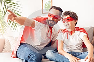 Father shwoing son view out the window dreamful in superheroe costumes at home sitting on sofa photo