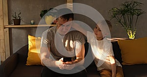Father sits on couch and plays in smartphone. Little son next to him interferes with his father and asks for attention