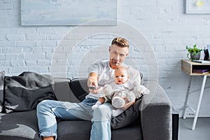 father sitting on couch with adorable baby daughter, holding remote controller and watching television