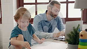 Father sitting at work using tablet while son drawing and painting, family relations concept