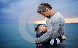 Father with small son on a walk outdoors standing on beach at dusk.