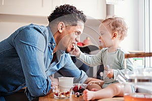 A father and a small toddler son eating fruit and yoghurt indoors at home.