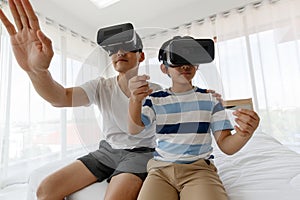 A father and son of an Asian family have fun and enjoy playing the game by wearing virtual glasses. On the bed in the room.