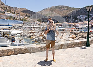 Father and son on the background of the port in the Greek city. The boy sits on dad& x27;s shoulders.