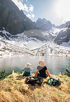 Otec a syn baťůžkáři odpočívají u horského jezera Zelené Pleso na Slovensku a užívají si zasněžené vrcholky. Jarní-letní turistika