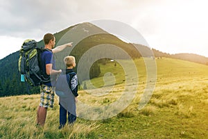 Father and son with backpacks hiking together in summer mountains. Back view of dad and child holding hands on landscape mountain