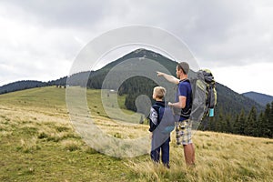 Father and son with backpacks hiking together in summer mountains. Back view of dad and child holding hands on landscape mountain