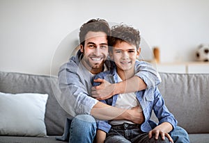 Father and son bonding. Happy dad embracing boy, posing and smiling to camera, sitting on couch at home