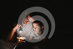 Father and son with flashlight reading book under blanket at home.