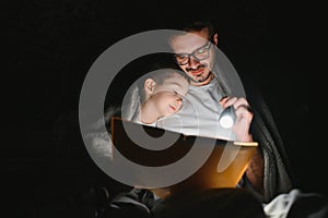 Father and son with flashlight reading book under blanket at home.