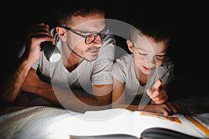 Father and son with flashlight reading book under blanket at home.