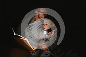 Father and son with flashlight reading book under blanket at home.