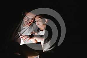 Father and son with flashlight reading book under blanket at home.