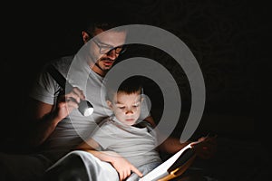 Father and son with flashlight reading book under blanket at home.