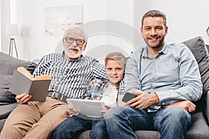 Father, son and grandfather relaxing together on couch in living room with digital tablet, smartphone