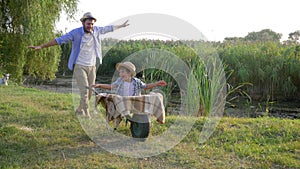 Father and son have fun with hands up, parent tickles little boy sitting in a wheelbarrow at village