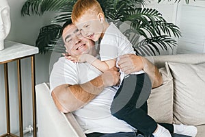 Father and son at home on the couch. Dad hugs his red-haired son tightly.