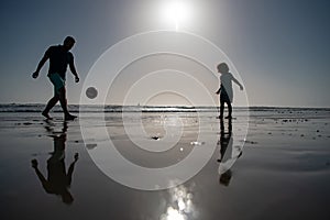 Father and son play soccer or football on the beach, silhouette on sunset. Dad and child having fun outdoors.